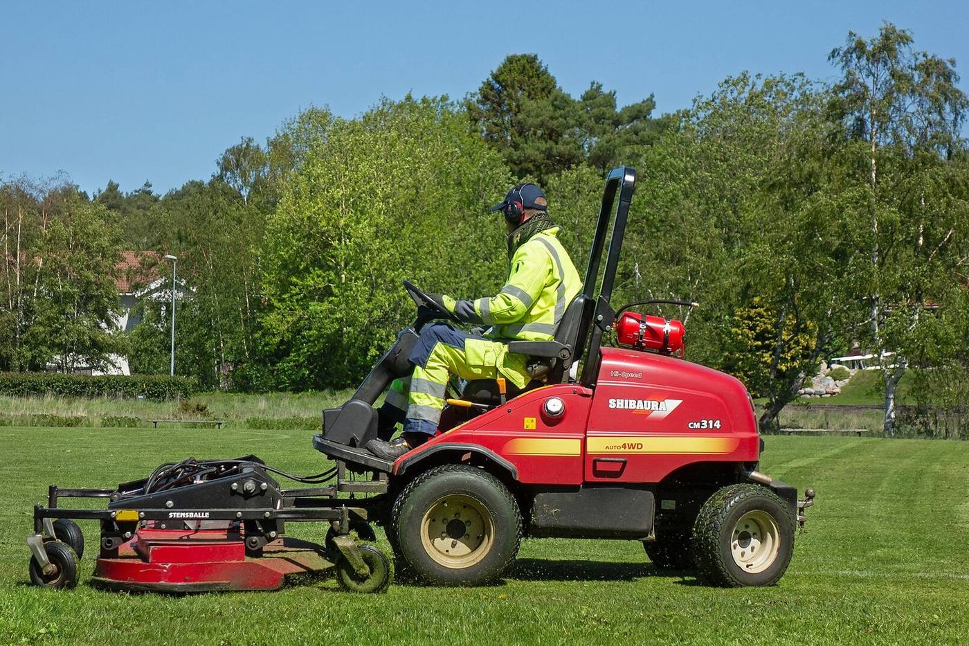A groundsman in a high-visibility jacket cutting a large grass field on a red ride-on mower, with trees behind