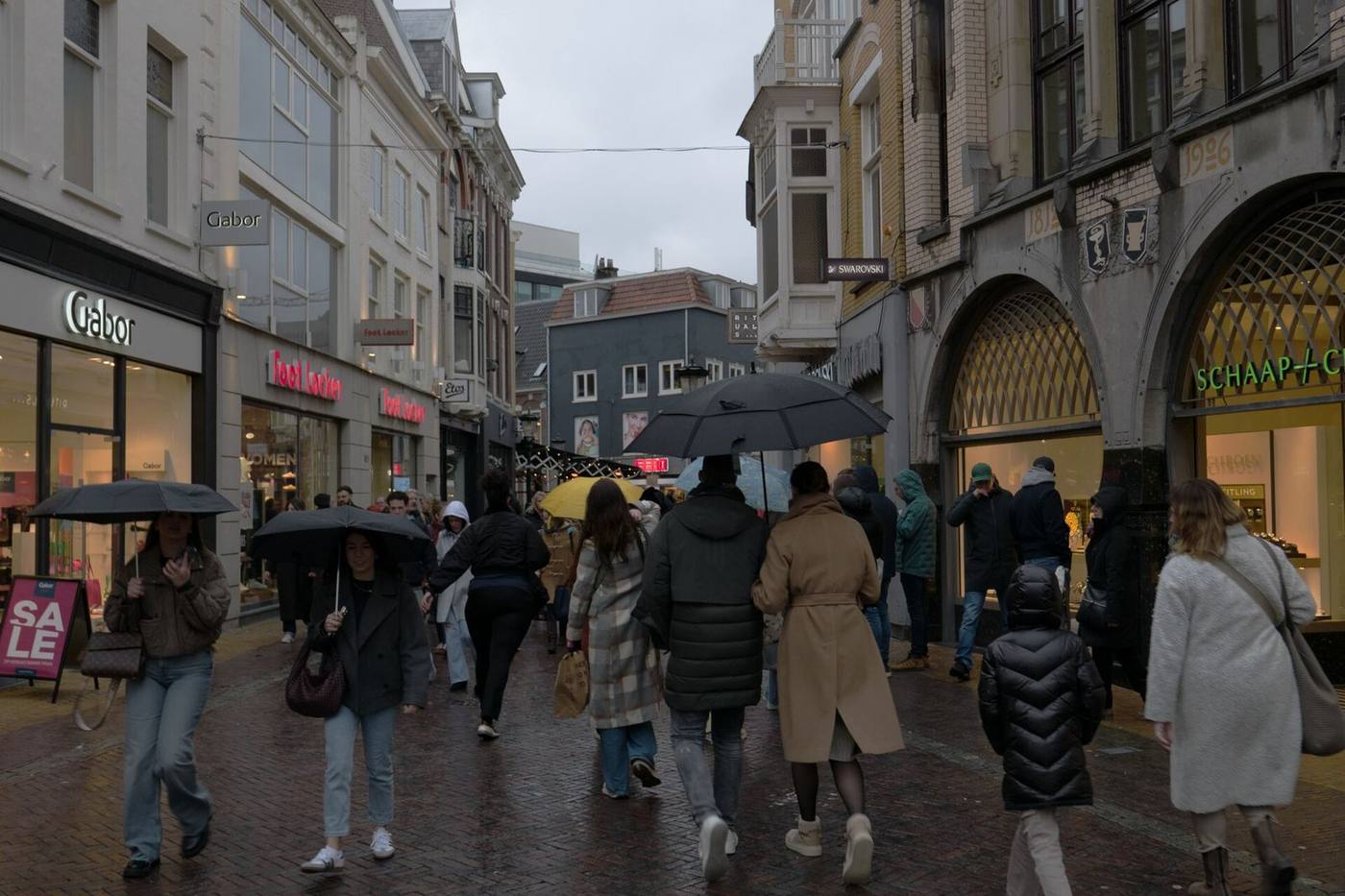 Shoppers with umbrellas walking along a wet pedestrian shopping street lined with lit shopfronts
